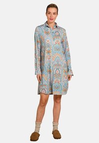 Vrouw in een langemouwen, gemêleerd blauw en beige hemdjurk met beige sokken en bruine pantoffels, staand tegen een witte achtergrond.