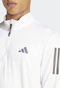 Biała polarowa bluza sportowa z kołnierzem na pół zamek, z szarym logo i trzema ciemnoszarymi paskami na lewym rękawie. Gładki, lekki materiał.