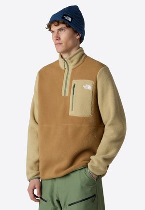 Pull en polaire beige et tan avec une fermeture éclair quart et une poche poitrine. Inclut un bonnet bleu côtelé et un pantalon vert, mettant en avant un design outdoor.