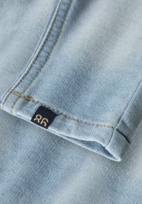 Tela de mezclilla azul claro con una textura distintiva, mostrando un borde doblado y costuras en color marrón. Se puede ver una pequeña etiqueta negra con "96".