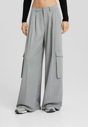 Woman wearing wide-leg gray pinstripe cargo pants with large side pockets and sneakers, standing against a plain background.