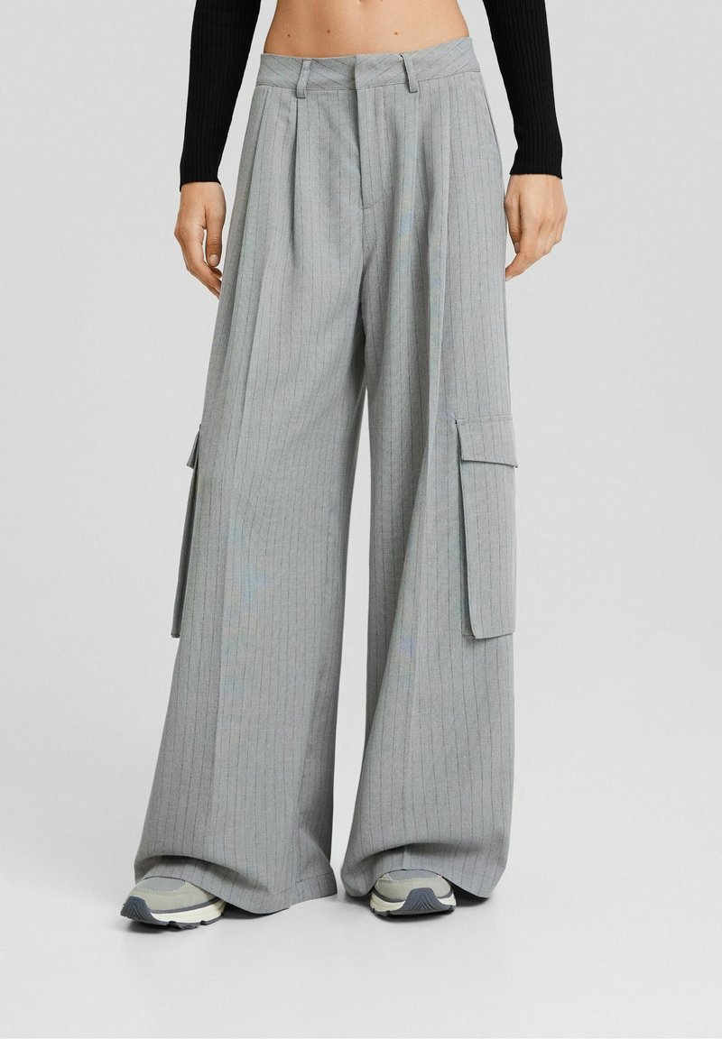 Femme portant un pantalon cargo gris à jambes larges avec fines rayures, grandes poches latérales et des baskets, debout devant un fond uni.