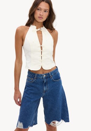 Jeune femme portant un top blanc sans manches à nouer sur le devant et un short bleu en jean ample avec des ourlets effilochés.