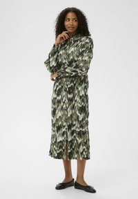 Rochie lungă cu model verde, cu fermoar în față, manșete strânse și fante laterale. Asortată cu pantofi negri cu talpă joasă. Textură netedă a țesăturii.