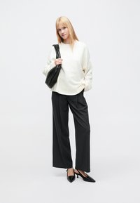 Kremfarget strikket genser, løs passform, ribbestrikket kant; mørke vide bukser; spisse svarte hæler; svart skulderveske; nøytral bakgrunn.