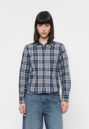 Young woman standing facing forward wearing a blue and white plaid button-up shirt and loose blue jeans against a plain background.
