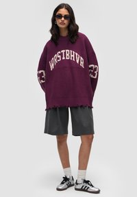 Übergroßer lila Strickpullover mit weißen Buchstaben "WRSTBHVA" und Akzenten von "23". Kombiniere ihn mit grauen Shorts und silbernen Sneakers. Mit ausgefranstem Saumdetail.