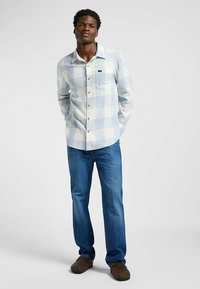Camisa de cuadros abotonada en azul claro y blanco, con un bolsillo en el pecho y botones marrones, combinada con vaqueros azules y zapatos marrones.