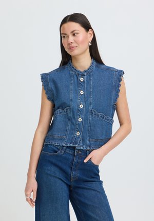 Ung kvinde med langt mørkt hår iført en ærmeløs denimvest med flæser og matchende højtaljede denimjeans, poserer med hånden i lommen.