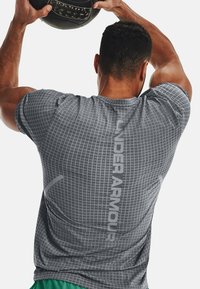 Šedé sportovní tričko s mřížkovou texturou a reflexní nášivkou "UNDER ARMOUR" na zadní straně, v kombinaci se zelenými šortkami, osoba držící fitness míč.