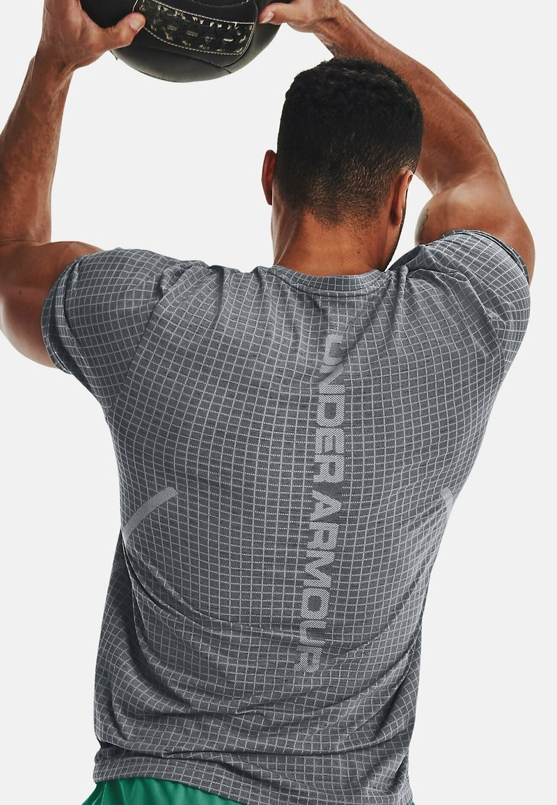 Šedé sportovní tričko s mřížkovou texturou a reflexní nášivkou "UNDER ARMOUR" na zadní straně, v kombinaci se zelenými šortkami, osoba držící fitness míč.