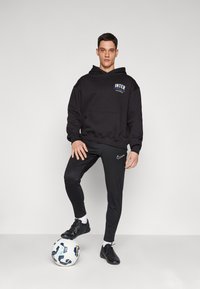 Hoodie preto com o logo "INTER", calças de desporto pretas e sapatilhas de futebol pretas. De pé sobre uma bola de futebol com padrão branco e azul.