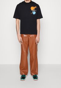 Black cotton T-shirt with vibrant orange and blue graphic on the chest, paired with brown faux leather wide-leg pants and green sneakers.