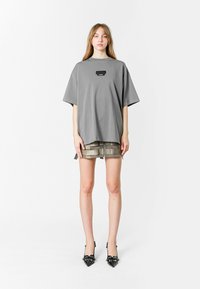 Camiseta gris de gran tamaño con una pequeña etiqueta negra en el pecho, combinada con pantalones cortos de textura metálica y tacones negros de punta.