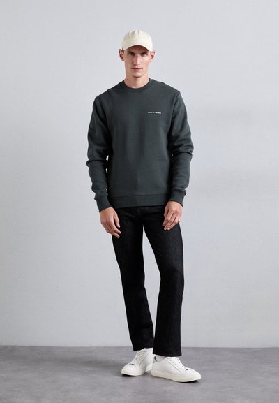 Dark green sweatshirt with a crew neckline and white text, paired with black jeans and white sneakers. Model stands against a light wall.