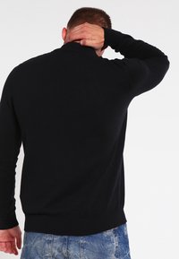 Svart tröja med rund halsringning, långa ärmar och ribbat avslut, bärs med blå denimjeans, sedd bakifrån. Slät textur.