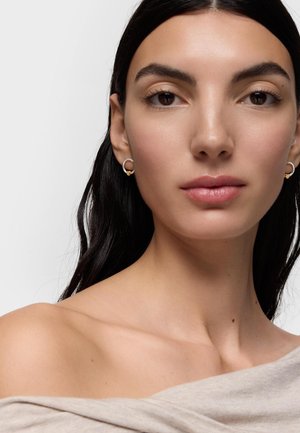 Mujer de cabello oscuro, con blusa beige con los hombros descubiertos y pendientes de aro plateados con detalles dorados, mirando hacia adelante sobre un fondo liso.