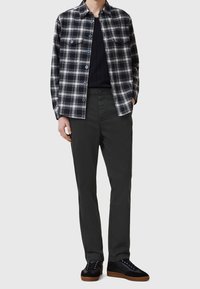 Osoba v černo-bílé kostkované košili s knoflíky přes černý top, tmavě šedých kalhotách a černých teniskách s hnědými podrážkami, stojící na bílém pozadí.