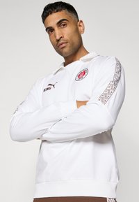 Witte polo met lange mouwen en een rits, met een cirkelvormig rood logo, bruine tekst "KING" en geprinte accenten op de mouwen.