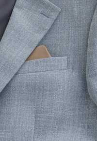 Blazer bleu clair texturé avec un mouchoir beige dans la poche poitrine sur une doublure intérieure foncée.