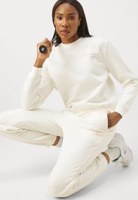 Felpa bianca in fleece e pantaloni jogger coordinati, con un sottile logo del marchio in pesca. Texture morbida con polsini elasticizzati e tasche.