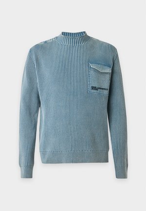 Homme debout portant un pull côtelé bleu avec poche poitrine, un jean bleu foncé et des baskets blanches sur un fond clair uni.