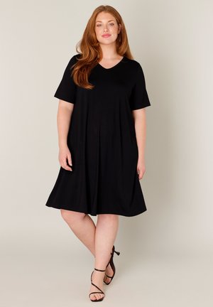 Vrouw met lang rood haar, gekleed in een zwarte jurk tot op de knie en zwarte sandalen met hoge hak en bandjes, staand tegen een effen achtergrond.