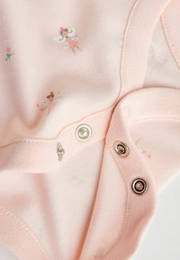 Šviesiai rožinė medvilninė kombinezono tipo palaidinė su fėjų ir gėlių raštu, apačioje su nikelio spaustukais ir minkštu, lygiu paviršiumi.