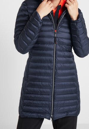 Osoba poprawiająca kołnierz ciemnoniebieskiej pikowanej kurtki puchowej sięgającej do kolan z przednim zamkiem błyskawicznym, noszonej na jasnopomarańczowej bluzce.