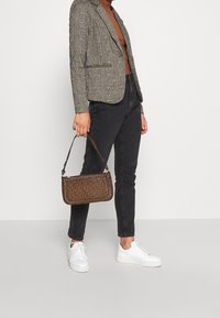 Brun mönstrad handväska med ett handtag upptill, bärs underarmen. Tyget har en texturerad yta. I kontrast, vita sneakers med en kraftig sula.