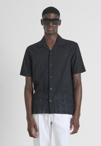 Camisa negra de manga corta con patrones de ojales, corte ligeramente holgado, con botones en el frente y cuello. Llevada con pantalones blancos.