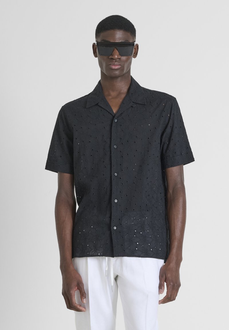 Zwarte shirt met korte mouwen met oogjespatronen, iets losse pasvorm, knoopsluiting aan de voorkant en een ceintuurneckline. Gekozen in combinatie met witte broek.