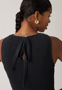 Νέα γυναίκα με πλεξούδα, που φορεί ασημένια σκουλαρίκια κρίκους και μαύρο υφασμάτινο αμάνικο τοπ με δέσιμο σε φιόγκο στο πίσω μέρος του λαιμού.