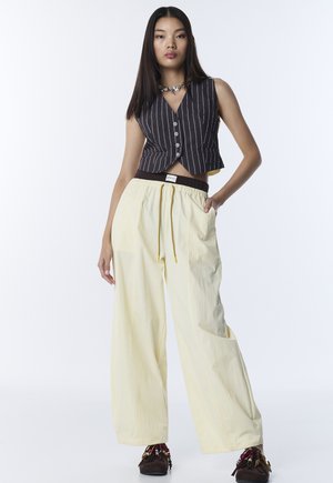 Femme aux longs cheveux foncés portant un gilet sans manches noir à fines rayures, un pantalon jaune clair à large jambe et des chaussures mocassins ornées de perles, debout avec une main dans la poche.