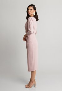 Jasnoróżowa średniej długości sukienka z rękawami trzyczwartowymi, dopasowany krój, gładka faktura i boczne rozcięcia. Noszona z cielistymi sandałami na wysokim obcasie.