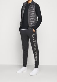 Pánský outfit se šedou péřovou vestou, černým dlouhým rukávem, tmavými joggery se znakem a bílými teniskami na jednobarevném pozadí.
