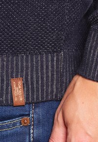 Granatowy sweter z dzianiny z teksturowanym wzorem, ściągaczami na mankietach oraz brązową skórzaną metką z nazwą marki. Widoczne dżinsy.