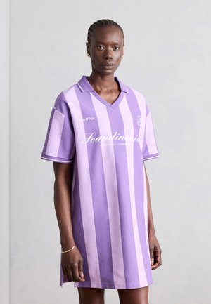 Kvinde med flettet hår iført en lilla og lavendelstribet polo kjole med teksten "Scandinavia Football Community", stående mod en ensfarvet baggrund.