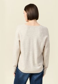 Vrouw met kort bruin haar, gekleed in een lichtbeige sweater en blauwe jeans, staand met de rug naar de camera tegen een effen achtergrond.