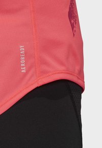 Gros plan sur un tissu de chemise de sport AEROREADY couleur corail rose sur un legging de sport noir avec des détails de couture visibles.
