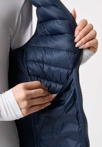 Hände, die eine marineblaue Steppjacke aufziehen und öffnen, um das Innenfutter und eine versteckte Tasche zu zeigen.