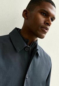 Primer plano de un hombre que lleva un abrigo abotonado gris oscuro y un suéter negro con cremallera, mirando ligeramente de lado contra un fondo claro.