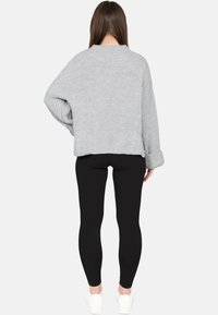 Donna in piedi di spalle che indossa un maglione in maglia grigio chiaro, leggings neri e sneakers bianche su uno sfondo semplice.