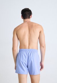 Lyseblå svømmeshorts med elastisk midje, laget av glatt stoff, vist fra baksiden på en bar overkropp mot en nøytral bakgrunn.