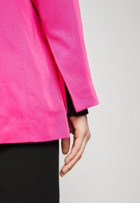 Osoba oblékající jasně růžový blazer s postranním rozparkem, černé kalhoty a černý rukáv viditelný pod rukávem blazera.