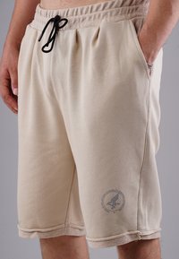 Beige cotton shorts with an elastic waistband, black drawstring, side pockets, and a logo detail on the left leg.