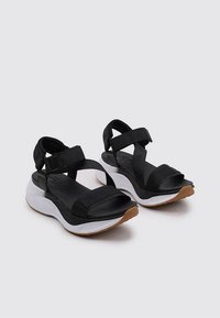Sandalias negras con correas ajustables, plantilla acolchada y suela de goma blanca y marrón. Diseñadas para mayor comodidad y uso diario.
