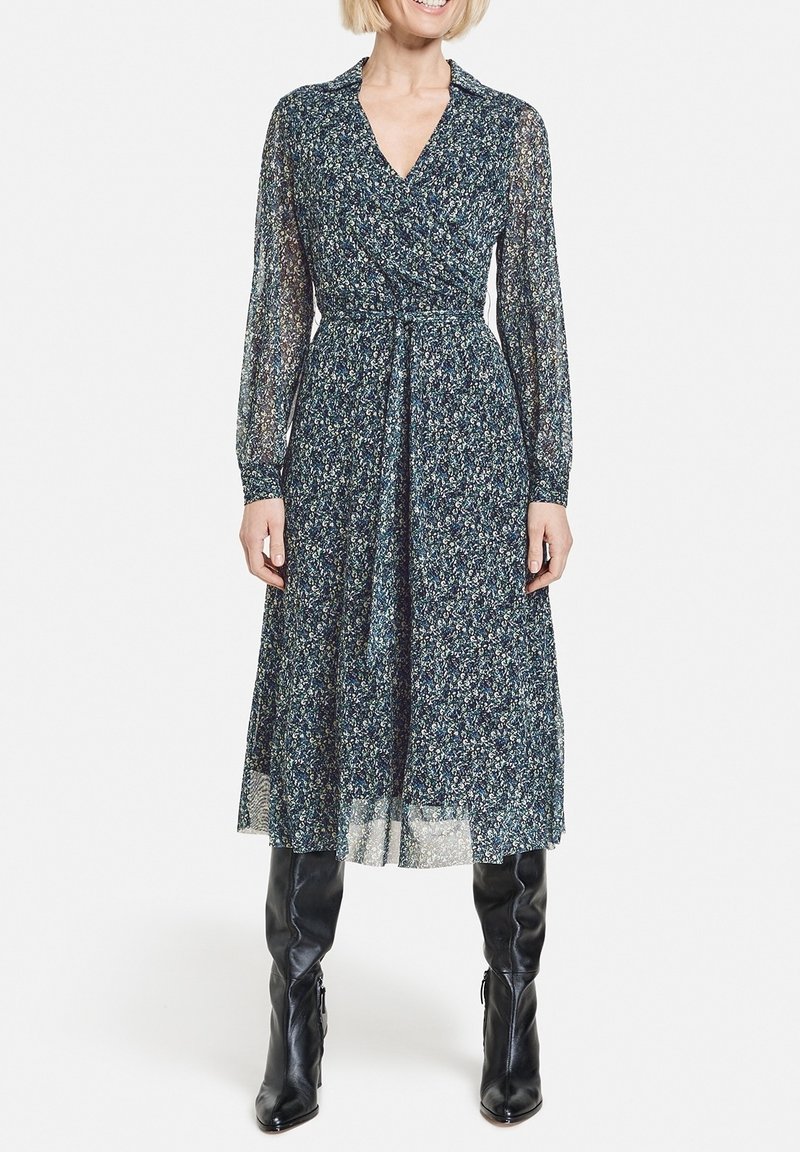 Femme portant une robe longue à manches longues, longueur cheville, bleue à motifs floraux, avec un décolleté en V et des bottes en cuir noires montantes jusqu'aux genoux, sur fond blanc.
