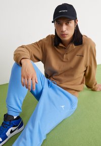 Bruine lange mouwen polo shirt met zwarte kraag, lichtblauwe sweatpants met een wit logo, zwarte pet en blauwe hoge sneakers, zittend op een groene vloer.