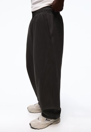 Mężczyzna w luźnych czarnych spodniach dresowych i beżowych butach do biegania, pokazany od talii do stóp na białym tle.
